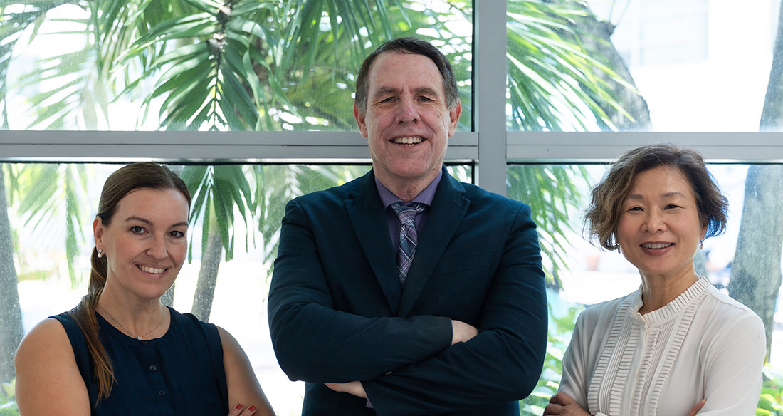 Dr. Rosie Curiel Cid, Dr. David Loewenstein, and Dr. Diane Zheng pose together indoors in front of large windows with palm trees visible outside.