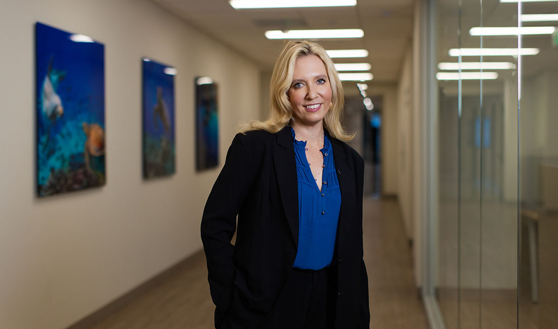 Dr. Tracy Crane stands confidently in a modern office hallway with glass walls and framed artwork, wearing a blue blouse and black blazer.