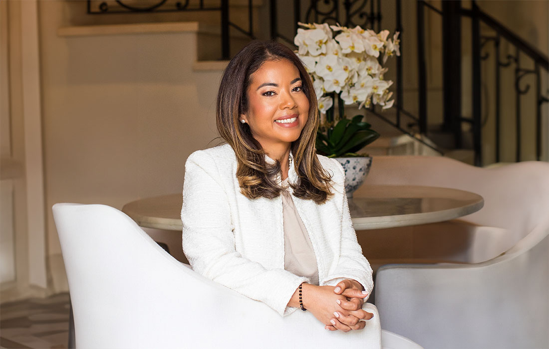Dr. Oriana Damas, sitting in a white chair in a living room with orchids behind her