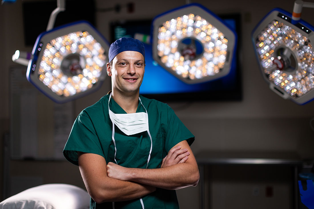 Dr. Ciro Amodio stands in an operating room wearing green surgical scrubs, a surgical cap, and a face mask around the neck. Illuminated surgical lights and medical equipment are visible in the background.