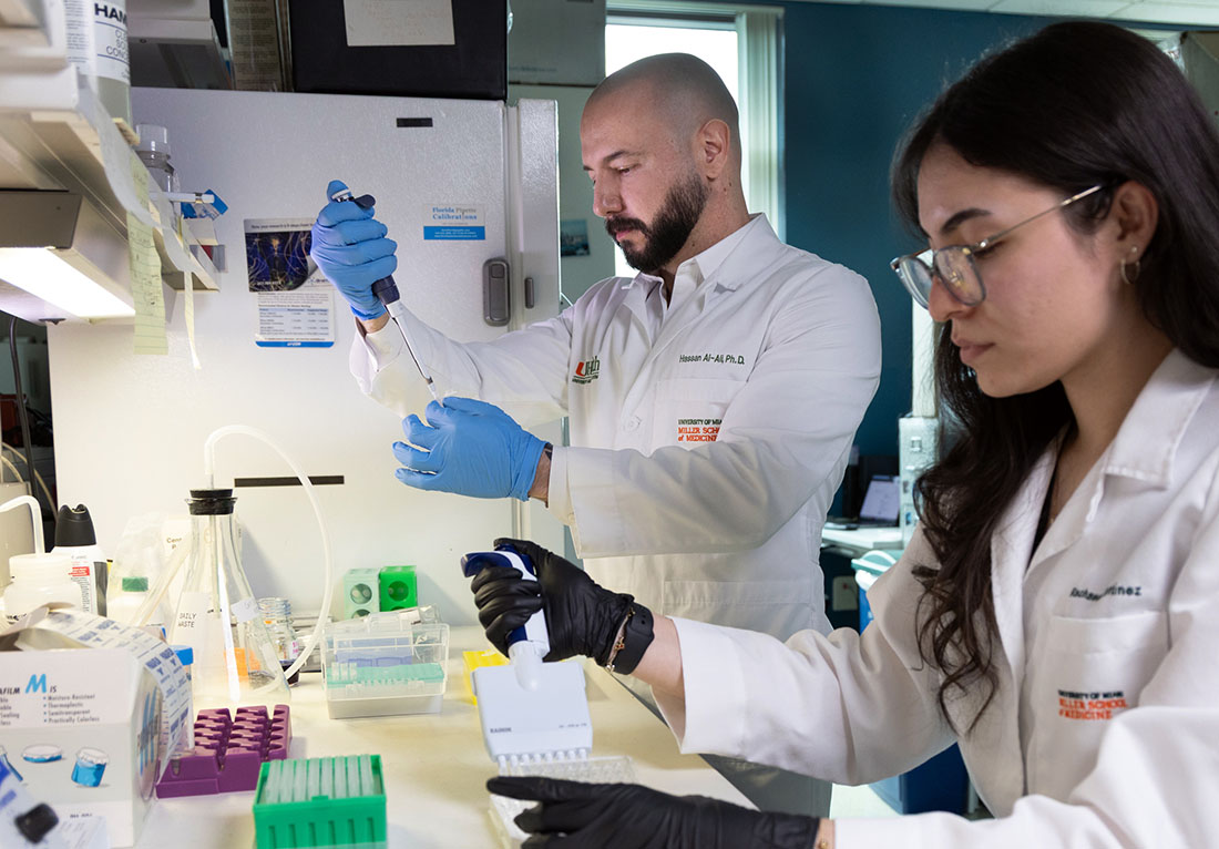 Dr. Hassan Ali, working in the lab with a colleague. Both are wearing white medical coats and manipulating lab equipment