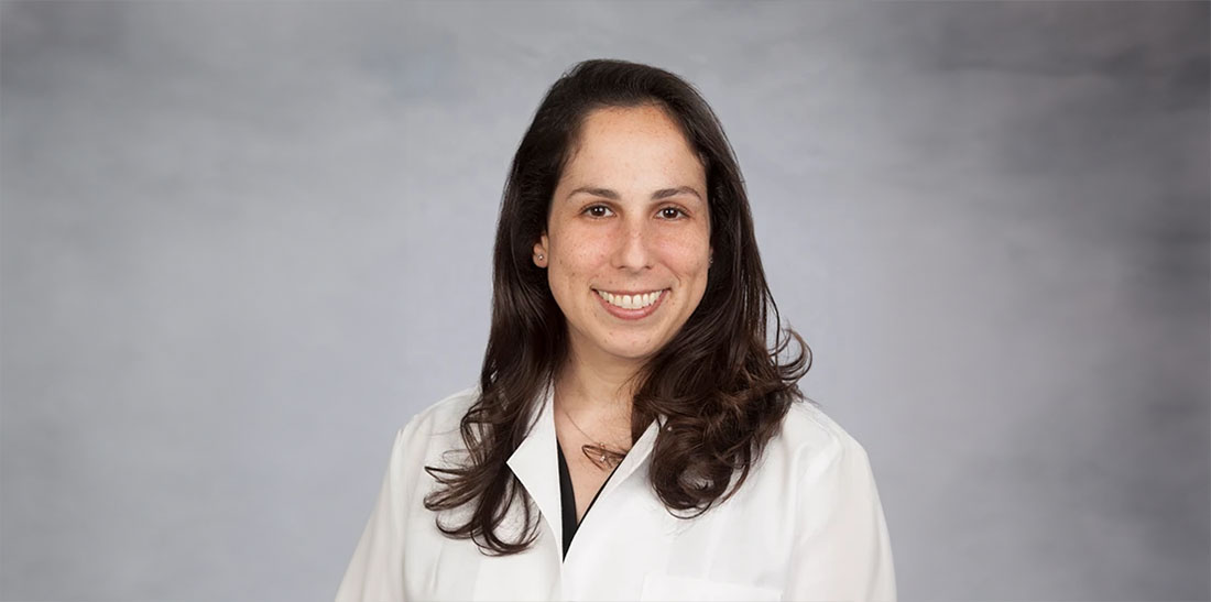 Studio portrait of Dr. Holly Cukier wearing a white lab coat, standing against a gray background and facing the camera.