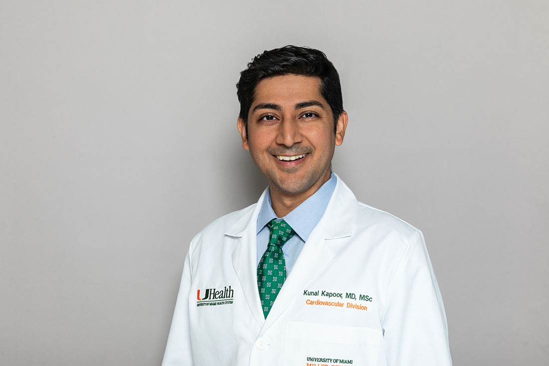 Portrait of a physician in a white UHealth lab coat, light blue dress shirt and green patterned tie standing against a light gray studio background. The embroidered lab coat identifies Kunal Kapoor, M.D., M.Sc., of the Cardiovascular Division.
