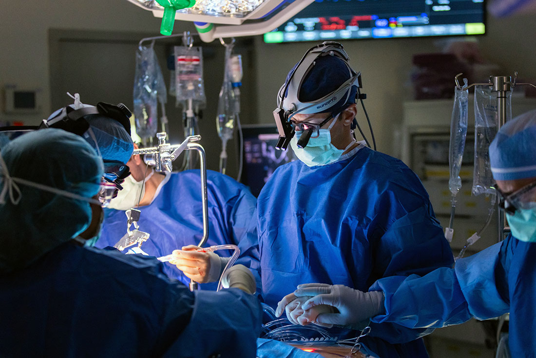 Three surgeons in full surgical attire work collaboratively over a patient during a cardiovascular procedure in a hospital operating room, with surgical instruments, drapes, and monitoring equipment visible.
