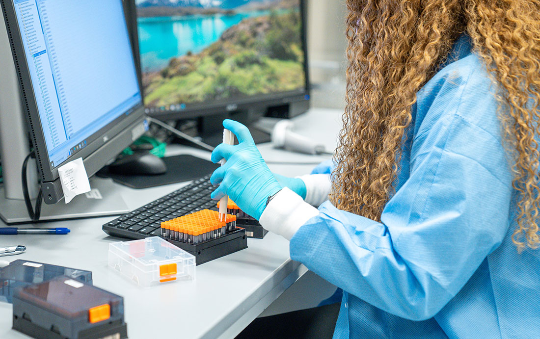 Laboratory researcher wearing gloves uses a pipette to transfer samples into a multiwell plate at a workstation with dual computer monitors in a scientific lab.