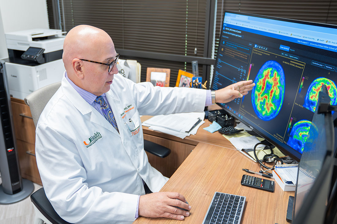 Dr. James Galvin in a white clinical coat sits at a desk reviewing colorful brain imaging scans displayed on a large computer monitor in an office setting.