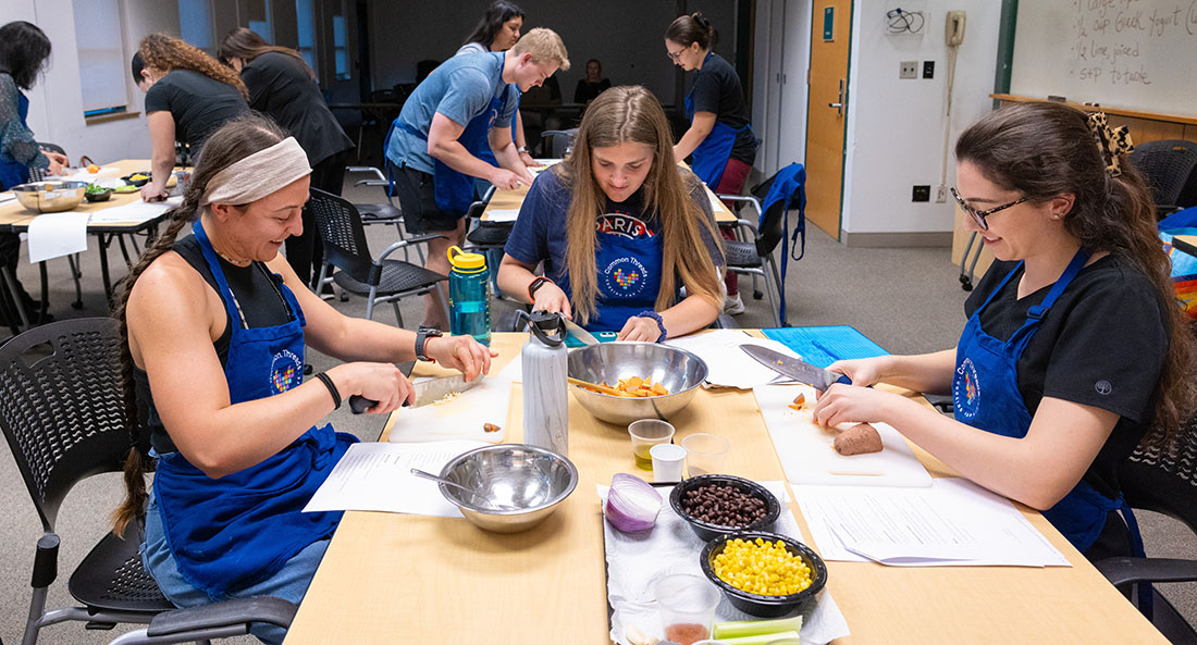 Miller School of Medicine students wearing blue aprons prepare food at tables in a classroom kitchen, chopping vegetables and mixing ingredients in bowls while following printed instructions, with additional participants working at stations in the background.