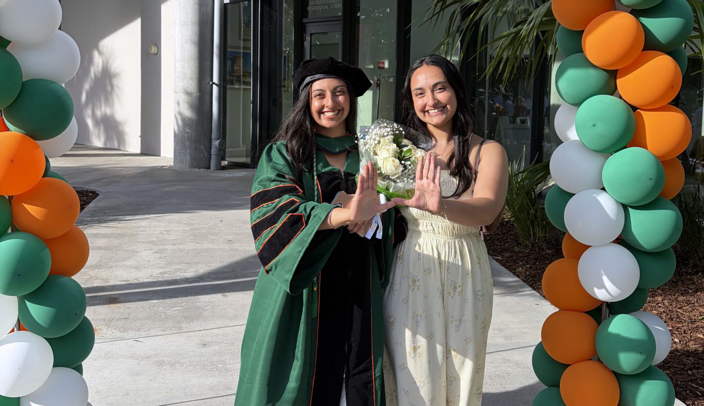 Serena Raj, a University of Miami Miller School of Medicine M.D./M.P.H. student, stands with her sister, Miller School graduate Aneesha Raj, M.D., M.P.H. ’26, beneath a University of Miami-themed balloon arch during a graduation celebration. Serena Raj wears academic regalia and holds a bouquet while both individuals form the University of Miami “U” hand gesture.