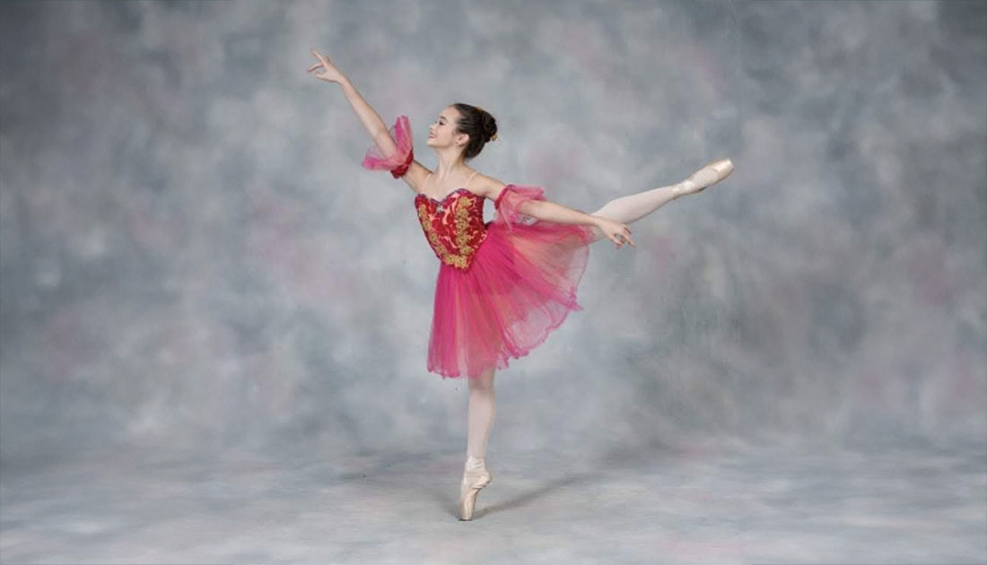 Nicole Kujas performs a ballet arabesque in a pink costume during a posed studio dance portrait against a neutral backdrop.