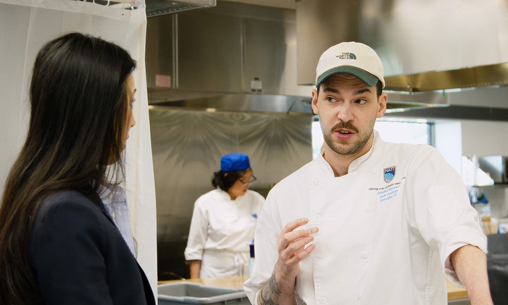 Person in a chef’s uniform and cap speaks with another person in a commercial kitchen, while a second chef works in the background near stainless steel equipment.