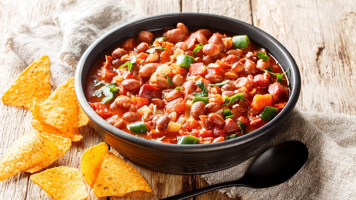 A bowl of stew with pinto beans and some chips beside it sits on a table.