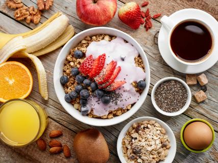 Bowl of granola and oats topped with yogurt and berries, with fresh fruit, coffee, eggs and seeds on table
