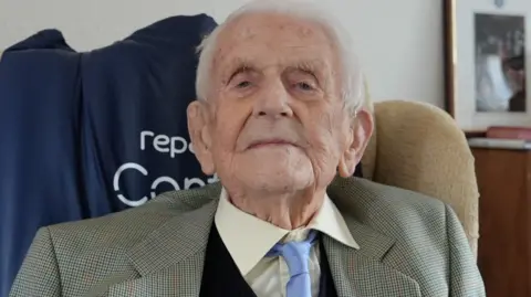Qays Najm/BBC Ron Letheren has white hair and is looking directly and slightly down at the camera. He is wearing a checked blazer, navy jumper, light blue tie and cream shirt. He is sitting in an armchair.