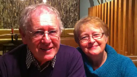 Keith Andrews Two adults sit side by side in a restaurant booth, leaning slightly towards one another as they face the camera. The person on the left, Keith, wears rectangular glasses, a dark purple jumper and a checked shirt, while the person on the right, his wife Sue, wears glasses and a bright blue knitted sweater. The image is closely framed around the two seated figures.