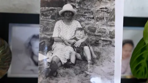 Qays Najm/BBC A black and white photo with Letheren as a toddler sitting on a beach against a rock wall, with a woman hugging him. She is wearing a hat and white patterned dress. They are both smiling widely at the camera.