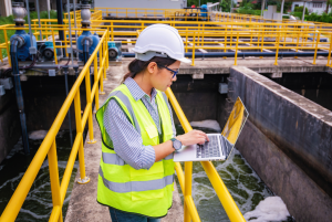 An engineer wearing a high-vis vest and hard hat inputs water quality data into a laptop at a wastewater treatment facility. Yellow safety railing is visible throughout the photo.