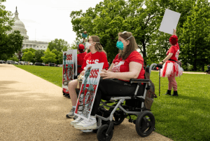 MEAction advocates demonstrate outside of the Capitol building in Washington D.C. on a sunny, spring day. They wear red t-shirts that read "millions missing" and "SOS" as well as high quality masks. One is in a wheelchair, while another sits on a stool. They hold signs that say, "Millions Missing, SOS, 2025, MEAction," that have an illustration of the Statue of Liberty.