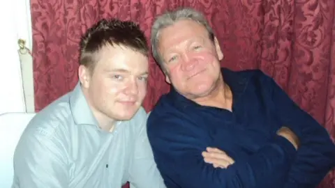 Michael Fathon A picture of a father and son. The younger man wears a pale grey shirt and the older man wears a navy shirt with his arms crossed. Both look relaxed and happy.