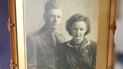 Qays Najm/BBC A black and white photo of Ron Letheren as a young man with thick, high hair. He is wearing a military uniform and is sitting close to a young woman with curled hair. She is wearing a collared dress with a brooch. They look serious.