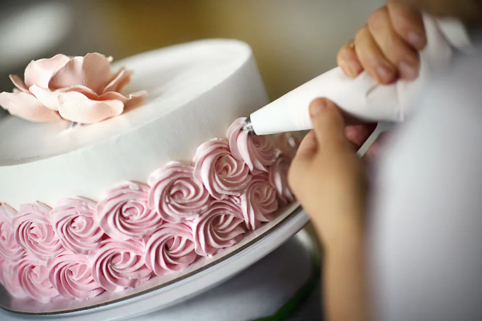 Person piping intricate pink rosettes on a cake topped with a single flower, showcasing detailed cake decorating skills