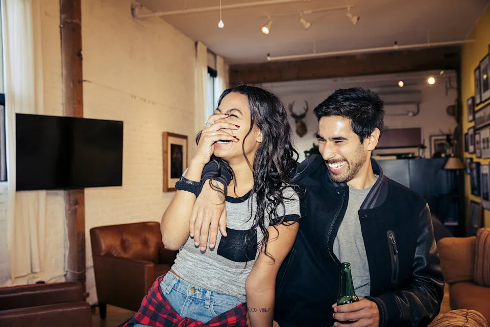 A couple laughs together in a cozy living room. The man holds a drink, and the woman covers her smile, showcasing a playful, intimate moment