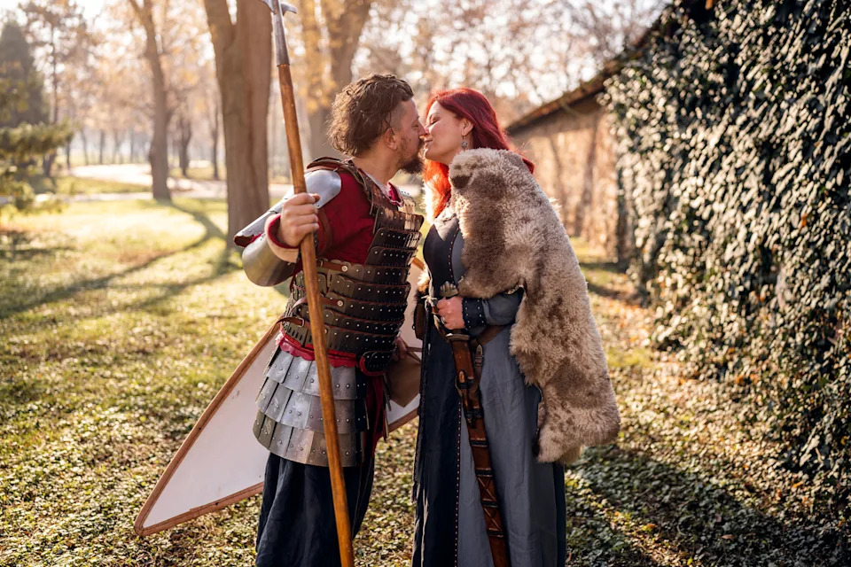 Two people in medieval-inspired outfits share a kiss outdoors. One holds a spear and a shield, while the other wears a fur cloak