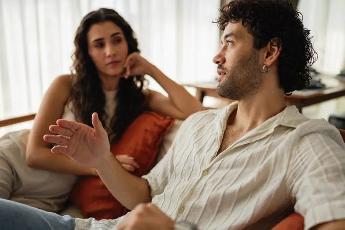 A couple sits together on a couch, engaging in a serious conversation. Both appear thoughtful and attentive to each other