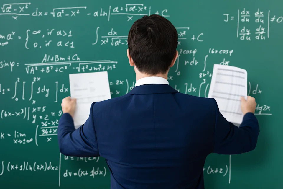 A person in a suit stands facing a chalkboard filled with complex math equations, holding papers, possibly solving problems