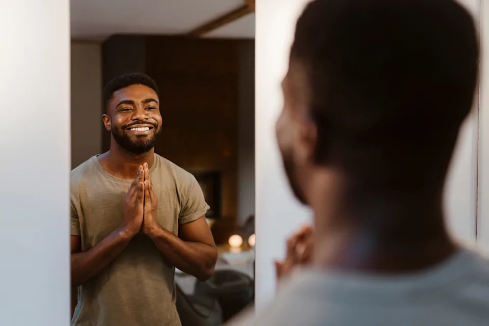 Man smiling at his reflection in a mirror, standing in a casual setting, hands clasped together