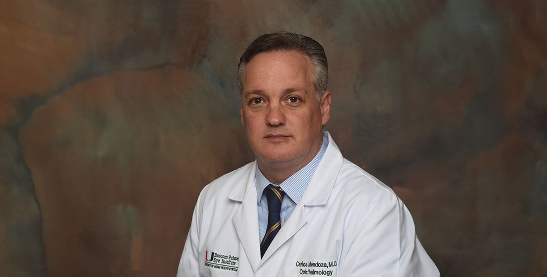 Carlos Mendoza-Santiesteban, M.D., poses for a professional portrait while wearing a white physician coat embroidered with the Bascom Palmer Eye Institute logo. The ophthalmologist is seated against a softly textured studio backdrop and faces the camera. A dress shirt and striped tie are visible beneath the coat. The image accompanies coverage related to clinical care, ophthalmology research, or vision science at the University of Miami Miller School of Medicine.