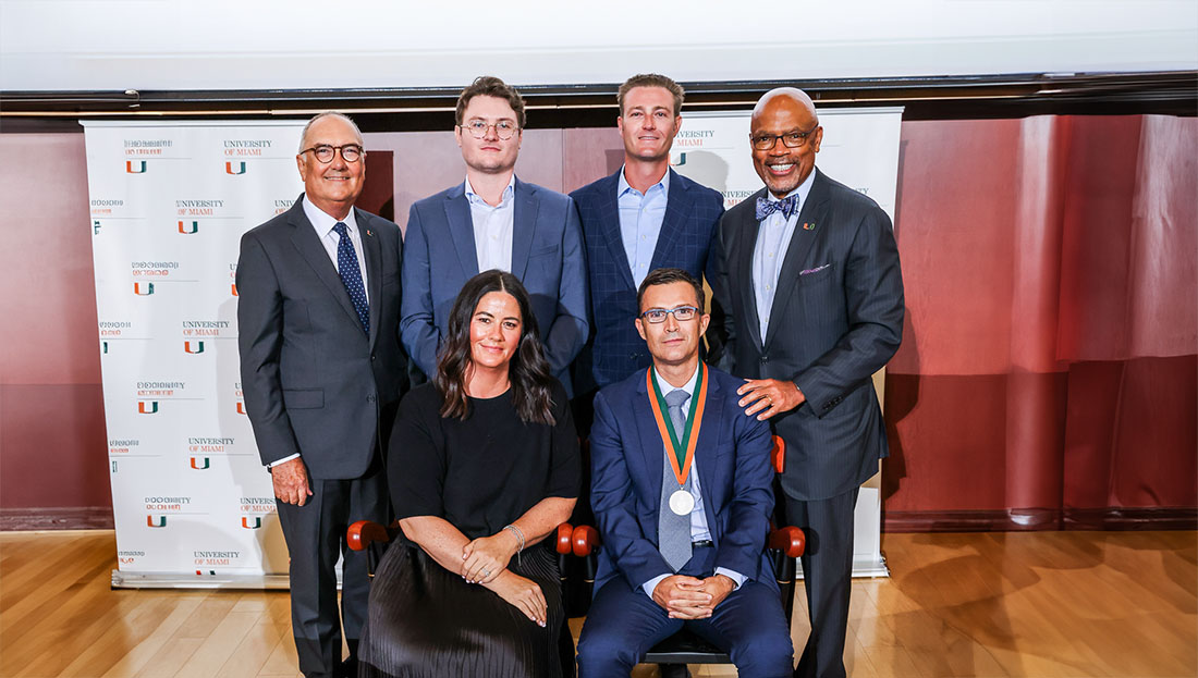 Kara Strobis Donvito, Guillermo Amescua, M.D., Eduardo Alfonso, M.D., Steffen Strobis, Peter Strobis and Dean Henri R. Ford pose for a group photograph at a University of Miami recognition event. One seated individual wears a ceremonial medal, while the group stands and sits in front of University of Miami-branded banners in an indoor auditorium setting.