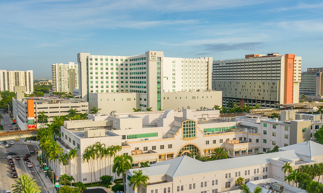 Aerial view of the UHealth medical campus in Miami, featuring University of Miami Health System hospital and clinical buildings, including the UHealth Tower, surrounded by palm trees, parking structures, and city streets under a clear blue sky.